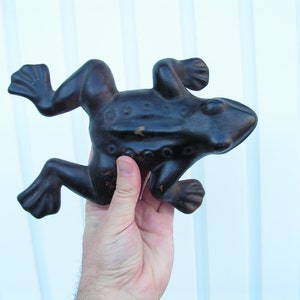 May include: A black cast iron frog doorstop with a textured surface. The frog is in a leaping pose with its legs outstretched.
