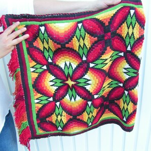 May include: A colorful, woven textile with a repeating floral pattern in red, orange, yellow, and green. The square-shaped piece has a black border with a green and red trim, and red fringe along the edges. The textile is held up by a person.