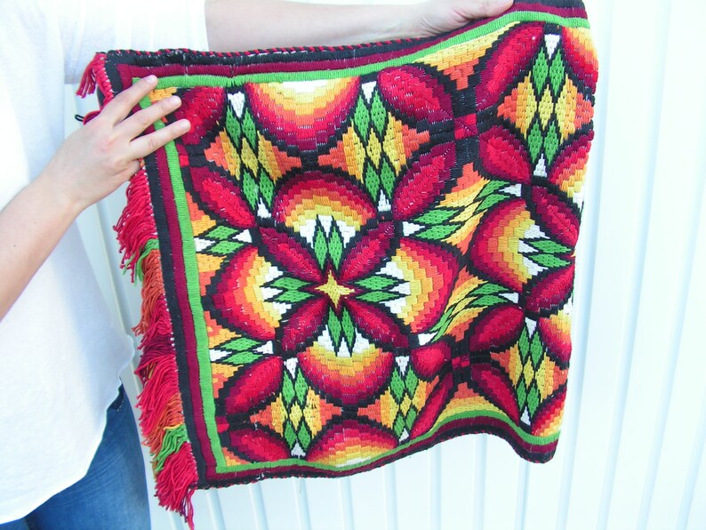 May include: A colorful, woven textile with a repeating floral pattern in red, orange, yellow, and green. The square-shaped piece has a black border with a green and red trim, and red fringe along the edges. The textile is held up by a person.