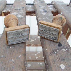 May include: Two vintage wooden stamps with text on the metal stamp face. The stamps have a rounded wooden handle and a rectangular metal plate with text. The stamps are resting on a weathered wooden surface.