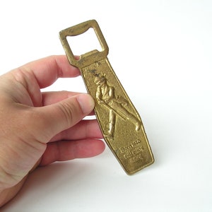 May include: A gold-coloured metal bottle opener with a raised design of a man in a top hat and a walking stick. The text "Born 1820 Still Going Strong 1963" is engraved on the opener.