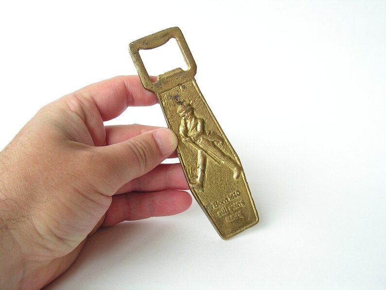 May include: A gold-coloured metal bottle opener with a raised design of a man in a top hat and a walking stick. The text "Born 1820 Still Going Strong 1963" is engraved on the opener.