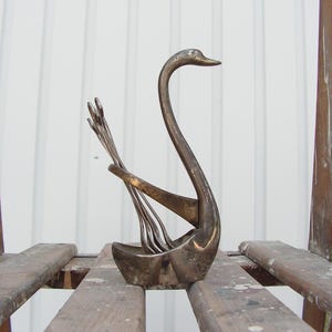 May include: A vintage silver swan-shaped utensil holder. The swan has a long, curved neck and holds several forks. The holder is made of metal and sits on a wooden surface. The background is a white wall.