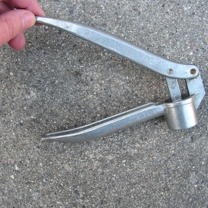 Könnte beinhalten: Silberfarbene Knoblauchpresse mit Scharnierdesign. Das Werkzeug verfügt über zwei gebogene Griffe und eine Presskammer. Die Presse ist aus Metall und für den Küchengebrauch konzipiert.