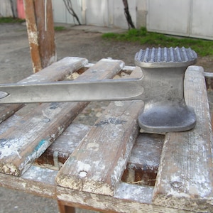 May include: A silver metal meat mallet with a textured head and a long handle. The mallet is resting on a wooden surface.