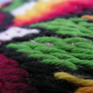 May include: Close-up of a colorful, textured textile. The image showcases a vibrant mix of colors, including green, black, red, yellow, and white. The woven pattern is visible, creating a tactile and visually appealing design. The textile appears to be handmade.