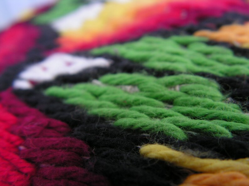 May include: Close-up of a colorful, textured textile. The image showcases a vibrant mix of colors, including green, black, red, yellow, and white. The woven pattern is visible, creating a tactile and visually appealing design. The textile appears to be handmade.