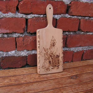 Op de afbeelding: Een houten snijplank met een decoratief ontwerp van een vrouw in een jurk en een kroon. Het ontwerp is in het hout gebrand.