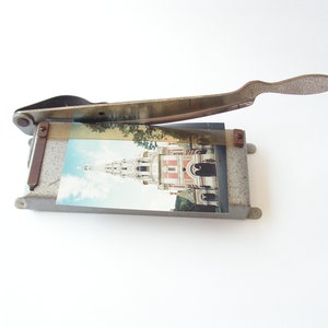 May include: A vintage metal paper cutter with a gray finish and a rusty handle. The cutter has a picture of a building with a spire on it.