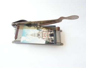May include: A vintage metal paper cutter with a gray finish and a rusty handle. The cutter has a picture of a building with a spire on it.