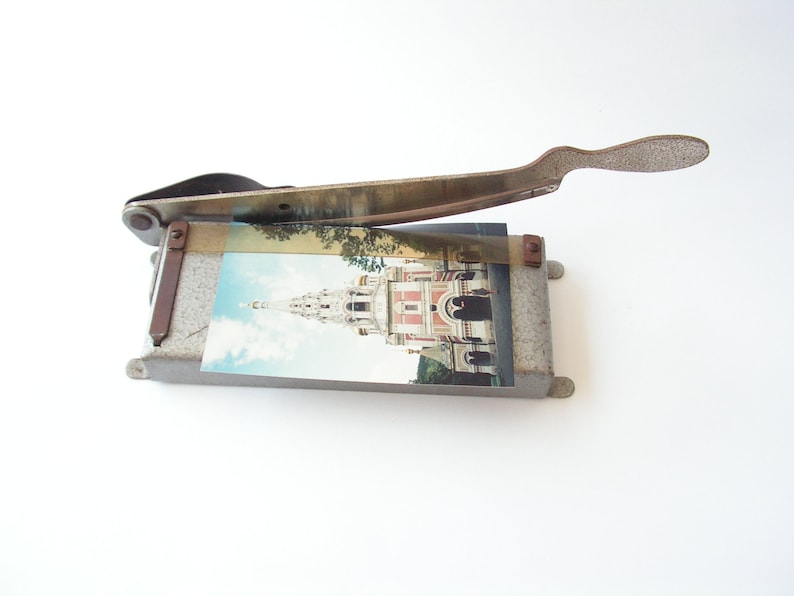 May include: A vintage metal paper cutter with a gray finish and a rusty handle. The cutter has a picture of a building with a spire on it.