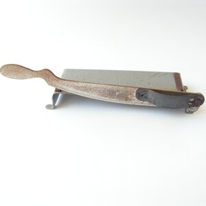 May include: A vintage metal paper cutter with a gray base and a rusty brown handle. The handle has a curved shape and a small metal loop at the end.
