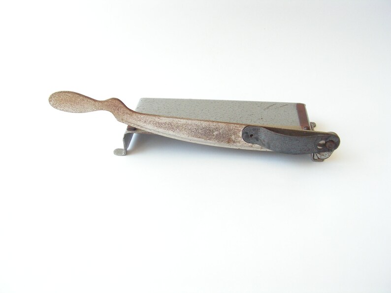 May include: A vintage metal paper cutter with a gray base and a rusty brown handle. The handle has a curved shape and a small metal loop at the end.