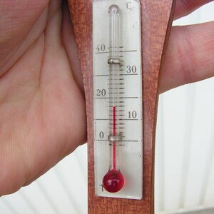 May include: A wooden thermometer with a red liquid inside. The thermometer is marked in Celsius and shows a temperature of 5 degrees.
