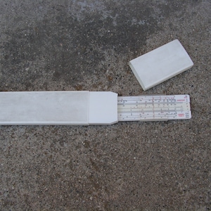 Könnte beinhalten: Ein weißes Plastik-Rechenschieber in einem weißen Plastik-Etui. Der Rechenschieber ist ausgezogen und das Etui ist geöffnet. Der Rechenschieber hat eine schwarze und rote Skala mit Zahlen und Markierungen.