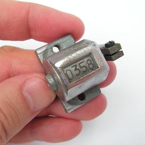 Op de afbeelding: Een kleine, vintage mechanische teller met een zilveren metalen behuizing. De teller geeft het nummer "0358" weer in een rechthoekig venster. Het apparaat heeft montagebeugels en een kleine hendel.
