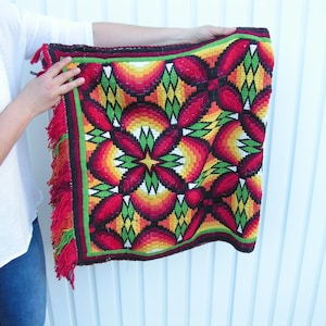 May include: A colorful, woven textile with a geometric floral pattern in red, orange, yellow, and green. The edges are trimmed with a green border and red fringe. The textile is held up by a person.