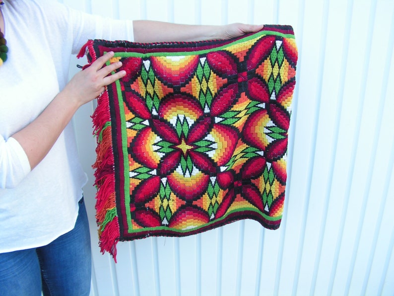 May include: A colorful, woven textile with a geometric floral pattern in red, orange, yellow, and green. The edges are trimmed with a green border and red fringe. The textile is held up by a person.