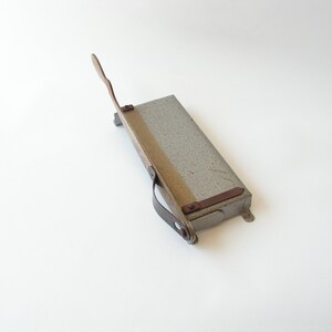 May include: A vintage metal paper cutter with a gray finish and a wooden handle. The cutter has a metal blade and a leather strap.