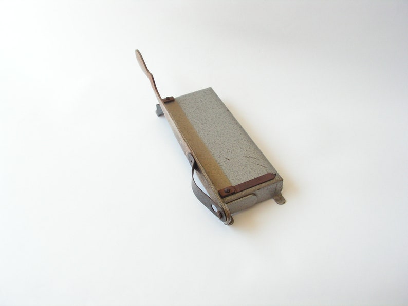 May include: A vintage metal paper cutter with a gray finish and a wooden handle. The cutter has a metal blade and a leather strap.
