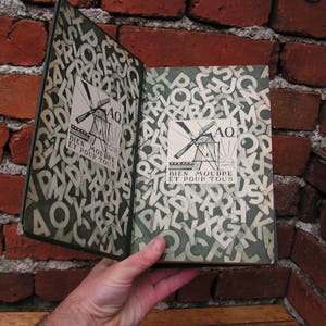 May include: An open book with a green cover and pages filled with white letters. The cover features a windmill illustration and the text "BIEN MOUDRE ET POUR TOUS". The book is held against a red brick wall.
