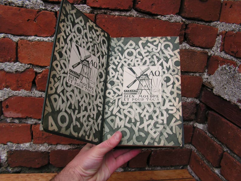 May include: An open book with a green cover and pages filled with white letters. The cover features a windmill illustration and the text "BIEN MOUDRE ET POUR TOUS". The book is held against a red brick wall.