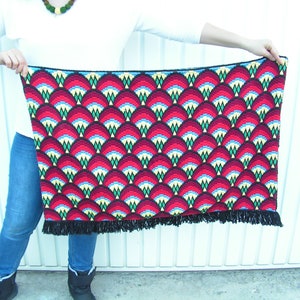 May include: A rectangular, patterned textile with a repeating fan-like design in red, black, blue, and yellow. The textile has a black fringe along the bottom edge. The textile is being held up by a person.