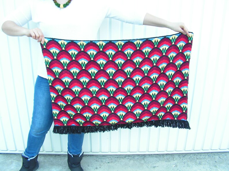 May include: A rectangular, patterned textile with a repeating fan-like design in red, black, blue, and yellow. The textile has a black fringe along the bottom edge. The textile is being held up by a person.