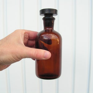 May include: A brown glass bottle with a black stopper. The bottle is empty and has a rounded shape.