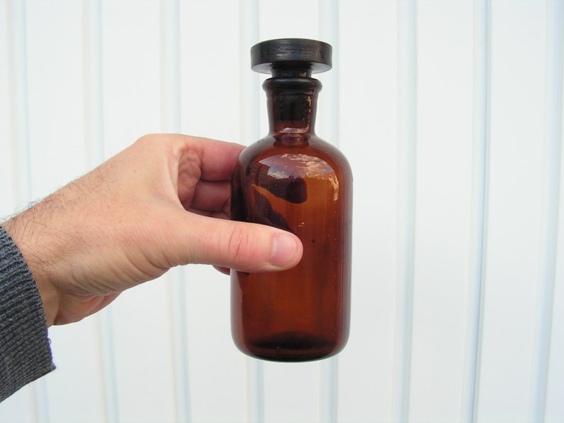 May include: A brown glass bottle with a black stopper. The bottle is empty and has a rounded shape.