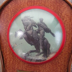 May include: A vintage wooden plaque with a red circular frame. Inside the frame is a picture of a person riding a horse. The picture is behind a clear plastic cover.