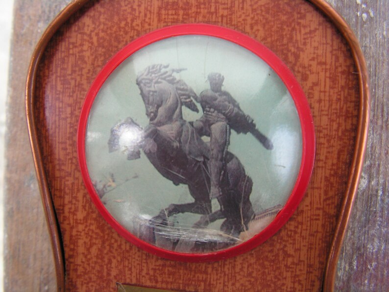 May include: A vintage wooden plaque with a red circular frame. Inside the frame is a picture of a person riding a horse. The picture is behind a clear plastic cover.