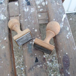 May include: Two wooden hand stamps with rounded handles and rectangular bases. Each stamp features a rubber printing surface attached to the bottom of the light brown wood block. The stamps are positioned on a weathered, paint-splattered wooden surface.