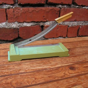 May include: A green paper cutter with a metal blade and a yellow handle. The cutter is sitting on a wooden surface.