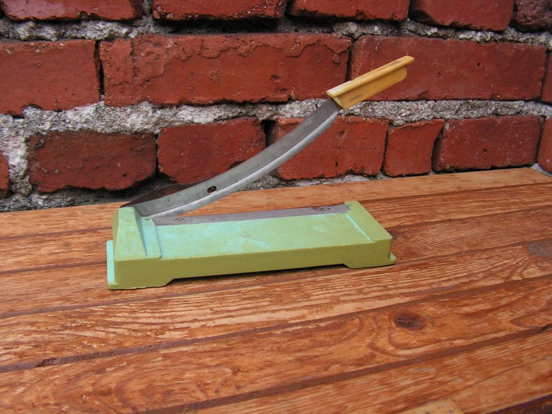 May include: A green paper cutter with a metal blade and a yellow handle. The cutter is sitting on a wooden surface.