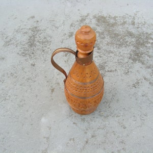 K&ouml;nnte beinhalten: Eine Holzflasche mit einem geschnitzten Design und einem Holzstopfen. Die Flasche ist braun und hat eine hellbraune Oberfl&auml;che. Die Flasche steht auf einer grauen Oberfl&auml;che.