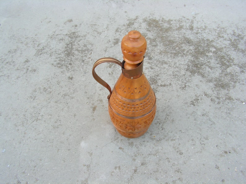 K&ouml;nnte beinhalten: Eine Holzflasche mit einem geschnitzten Design und einem Holzstopfen. Die Flasche ist braun und hat eine hellbraune Oberfl&auml;che. Die Flasche steht auf einer grauen Oberfl&auml;che.