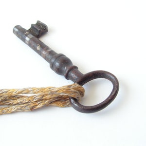 May include: An antique, rusty metal key with a circular handle and a textured shaft. The key is attached to a braided rope made of tan and gold threads. The key is set against a white background.