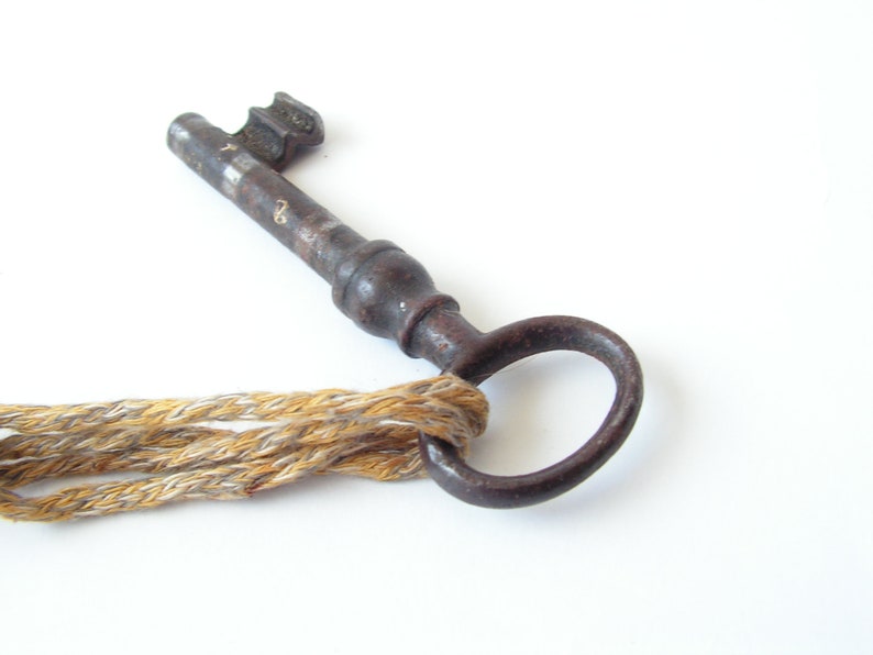 May include: An antique, rusty metal key with a circular handle and a textured shaft. The key is attached to a braided rope made of tan and gold threads. The key is set against a white background.