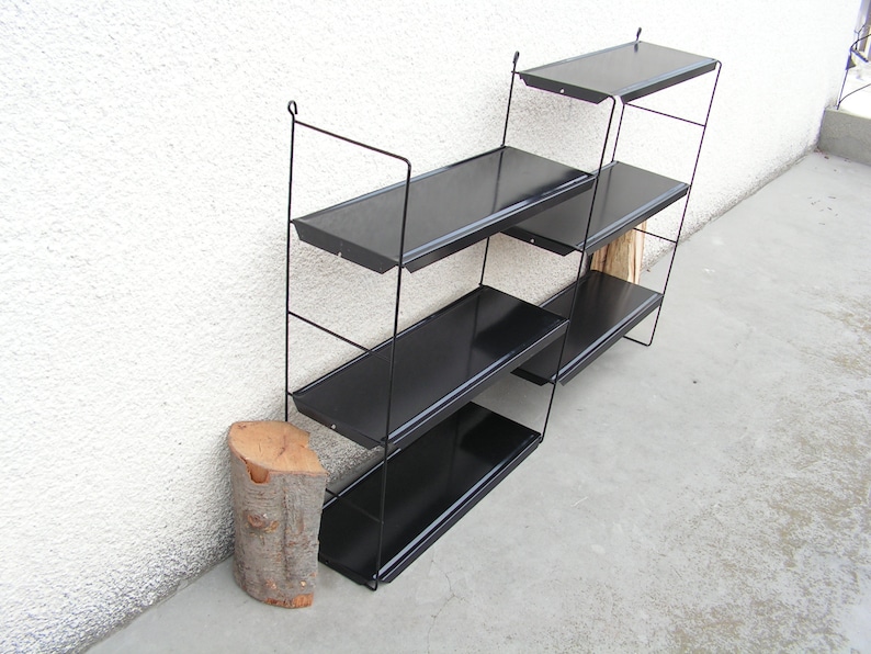 Puede incluir: Una estanter&iacute;a de pared de metal negro con cuatro estantes. Los estantes est&aacute;n inclinados y tienen un acabado negro. La estanter&iacute;a est&aacute; montada en una pared blanca.