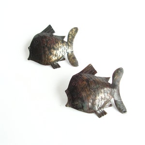 Può includere: Due pesci decorativi in metallo da appendere al muro. I pesci sono marroni e dorati con una superficie testurizzata. Entrambi sono rivolti verso il lato destro dell'immagine.