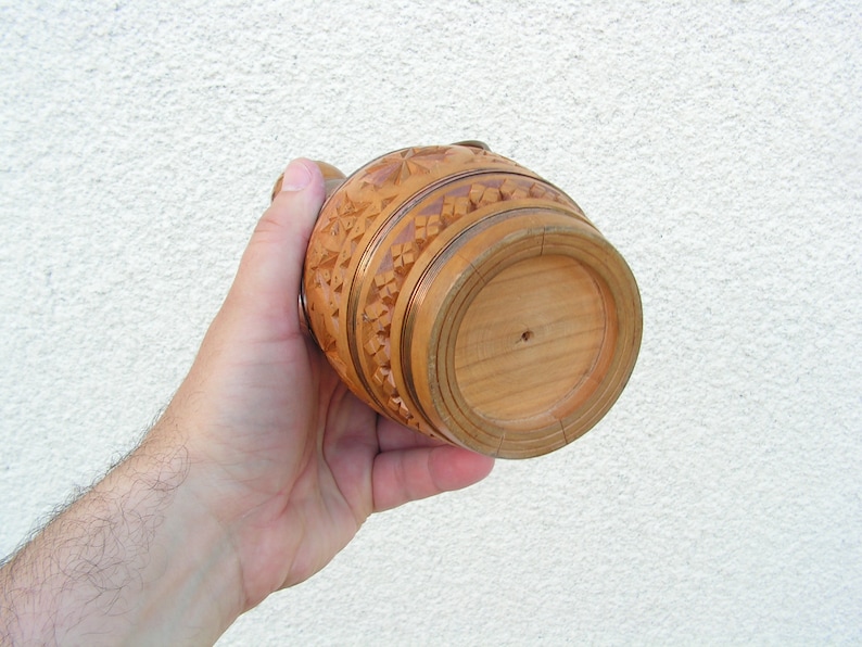 K&ouml;nnte beinhalten: Ein geschnitzter Krug aus Holz mit aufwendigen Details. Der Krug ist braun und hat eine runde Form. Der Boden des Kruges ist sichtbar.