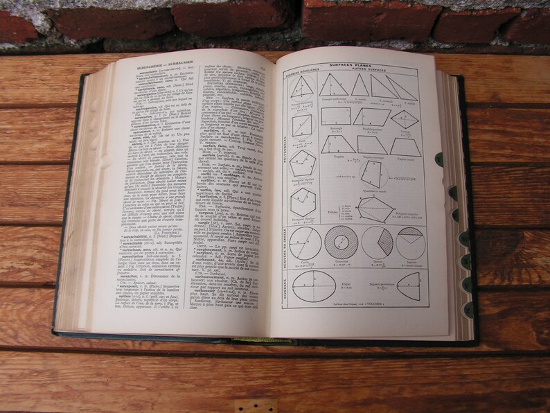 May include: An open vintage book with pages displaying text and geometric diagrams. The book is bound in a dark cover and rests on a wooden surface. The pages are aged with a yellowed hue, and the diagrams are in black ink.