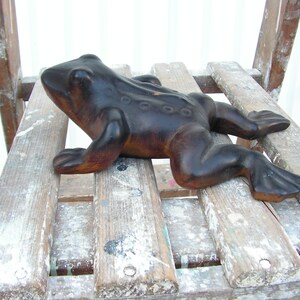 May include: A dark brown wooden frog figurine with a textured surface. The frog is lying on a wooden surface with white paint splatters.
