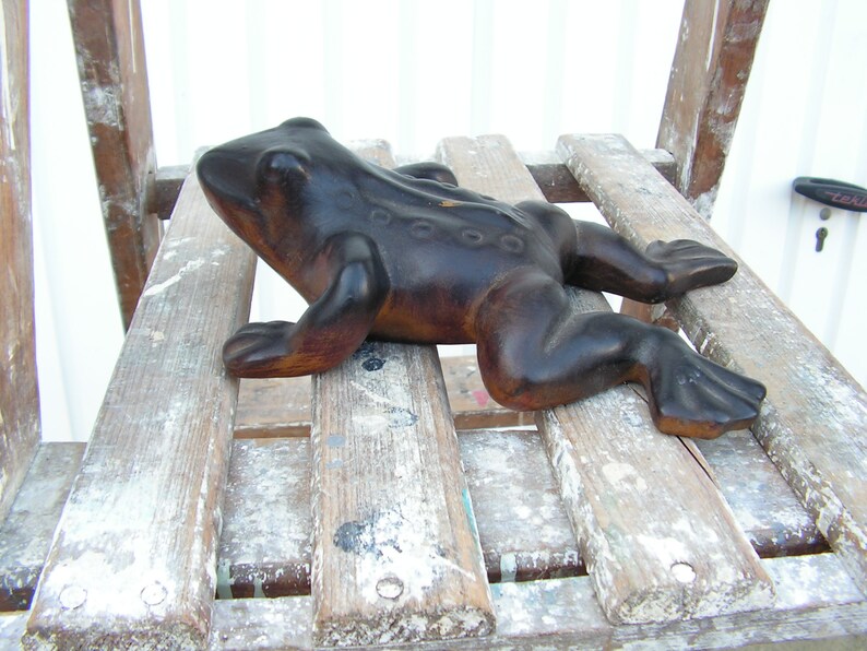 May include: A dark brown wooden frog figurine with a textured surface. The frog is lying on a wooden surface with white paint splatters.