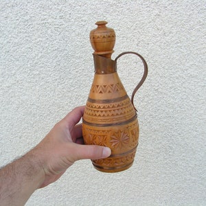 K&ouml;nnte beinhalten: Eine Holzflasche mit einem geschnitzten Design und einem kupfernen Deckel. Die Flasche hat einen Griff und ist aus hellem braunem Holz.