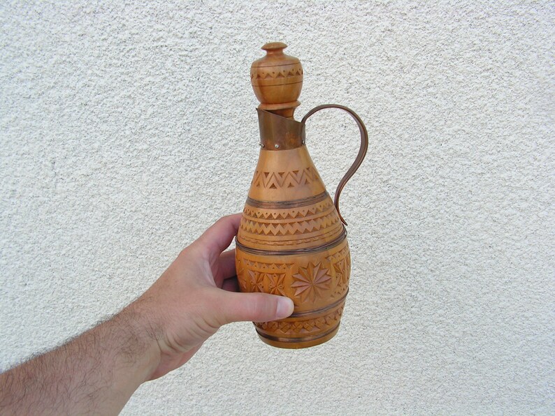 K&ouml;nnte beinhalten: Eine Holzflasche mit einem geschnitzten Design und einem kupfernen Deckel. Die Flasche hat einen Griff und ist aus hellem braunem Holz.