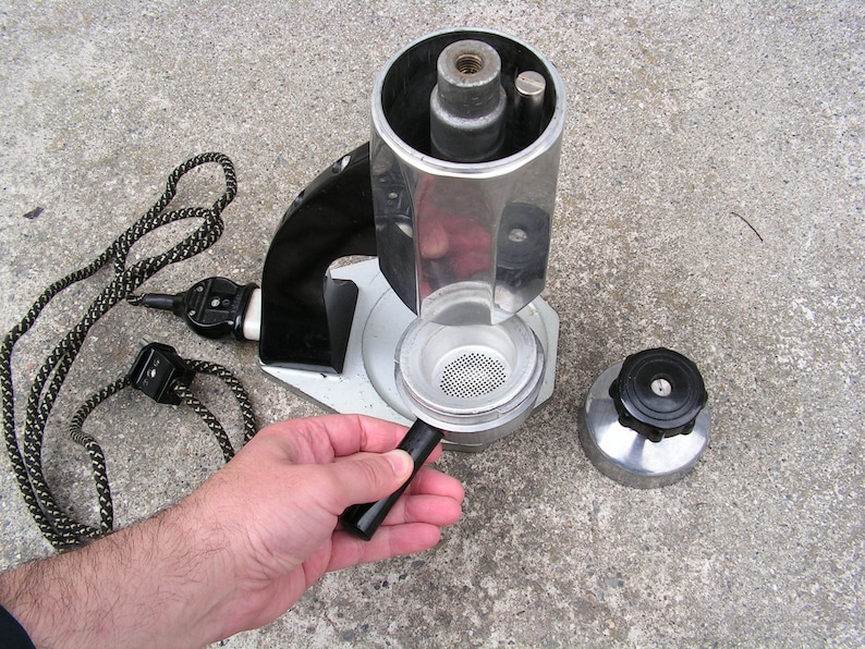 May include: A vintage silver and black electric coffee maker with a removable filter basket and a black handle. The coffee maker is disassembled and the parts are laid out on a gray concrete surface.