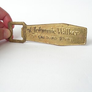 May include: A gold-coloured bottle opener with a rectangular opening and the words "Johnnie Walker Old Scotch Whisky" embossed on the top.