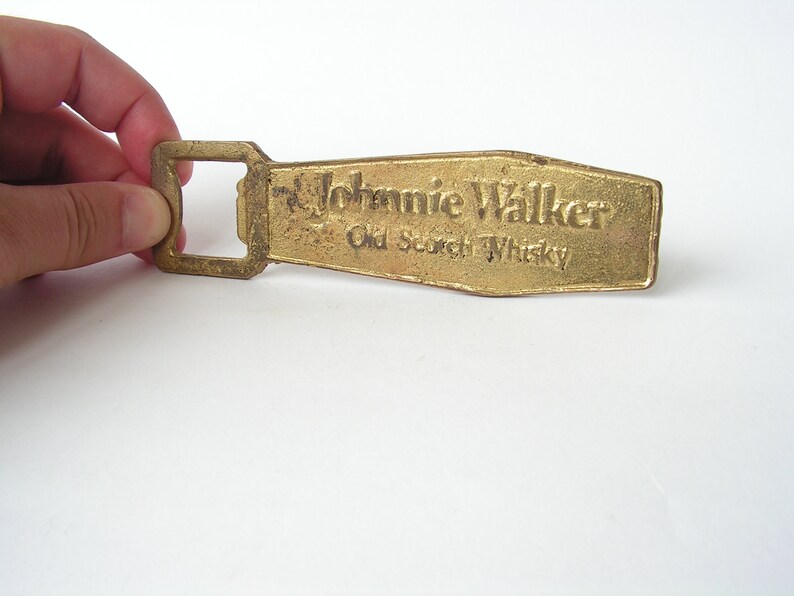 May include: A gold-coloured bottle opener with a rectangular opening and the words "Johnnie Walker Old Scotch Whisky" embossed on the top.